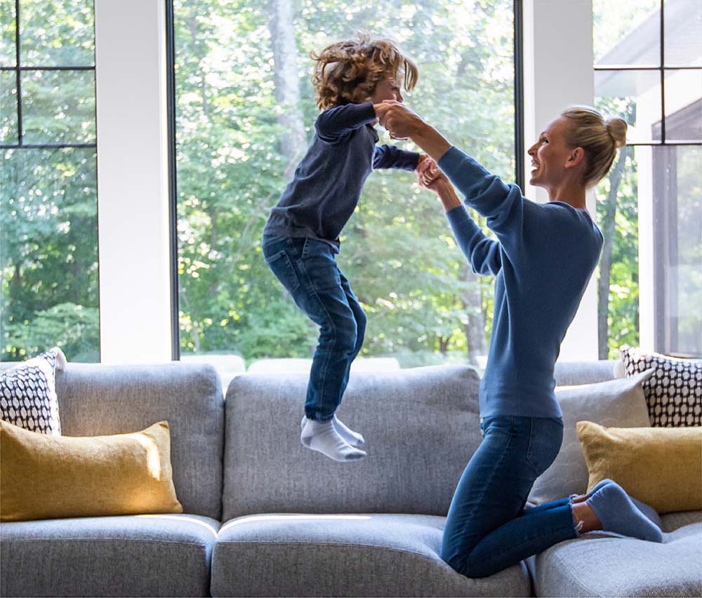 Child jumping on the couch holding his mom's hands.